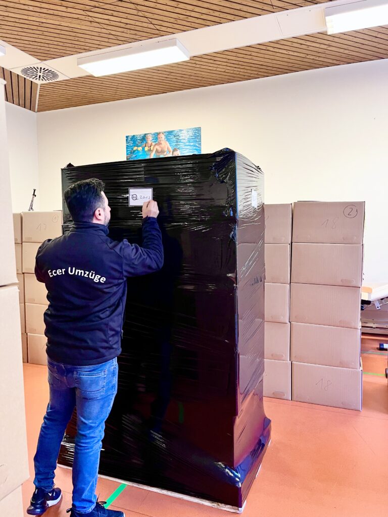 Mitarbeiter von Ecer Umzüge klebt ein Etikett auf ein in Folie verpacktes Möbelstück, umgeben von gestapelten Umzugskartons in einem Raum.