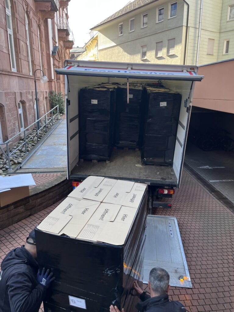 Geöffneter Umzugs-LKW mit im Inneren stehenden, schwarz in Folie verpackten Möbelstücken; im Vordergrund ein gestapelter Rollcontainer mit Umzugskartons, daneben die ausgefahrene Laderampe im Hofbereich.