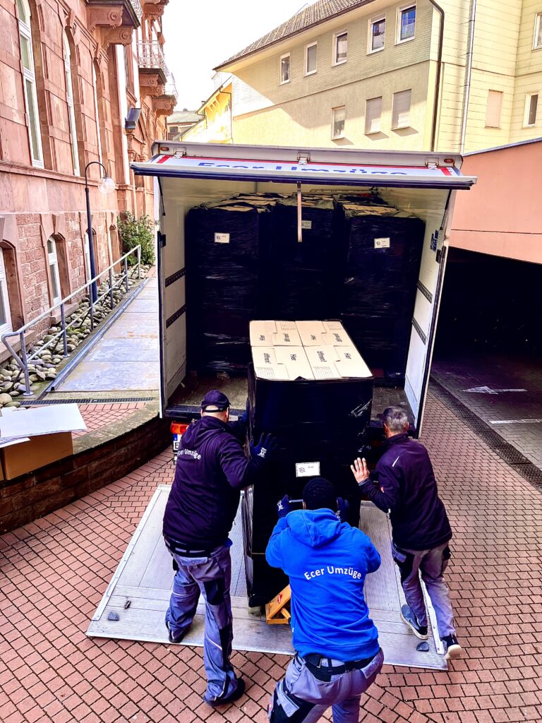 Zwei Umzugsmitarbeiter von Ecer Umzüge verladen ein großes, in Folie verpacktes Möbelstück aus einem geöffneten LKW in einer engen Hofeinfahrt.