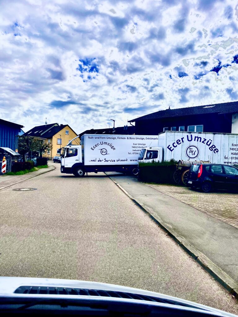 Zwei große LKW von Ecer Umzüge in einer Wohnstraße, einer im Abbiegevorgang, der andere seitlich mit großem Firmenlogo und Schriftzug „Ecer Umzüge …weil der Service stimmt“, daneben ein geparkter Pkw und ein alter Holzwagen.