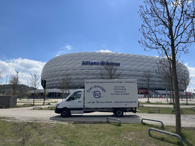 Transporter von Ecer Umzüge vor der Allianz Arena in München, geparkt an einer Straße mit Bäumen und blauem Himmel.