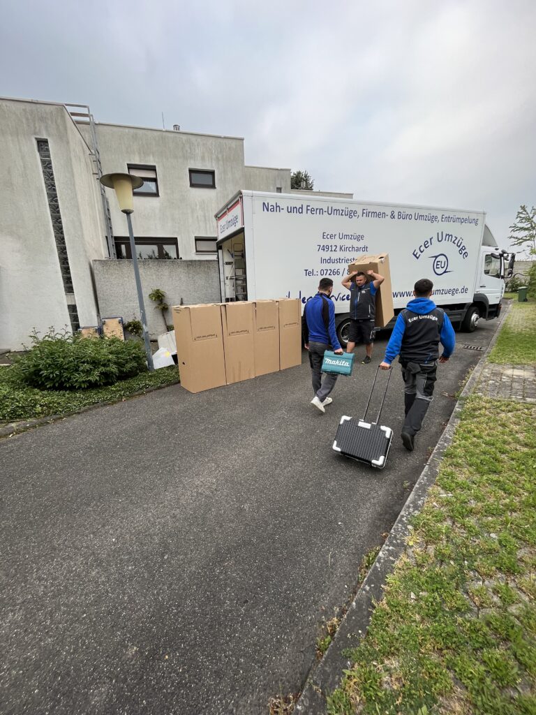 Drei Umzugshelfer von Ecer Umzüge transportieren Kartons, Werkzeugtasche und Rollbrett vom geparkten Umzugs-LKW in die Einfahrt eines Wohnhauses, neben einer Reihe aufgestellter Umzugskartons.